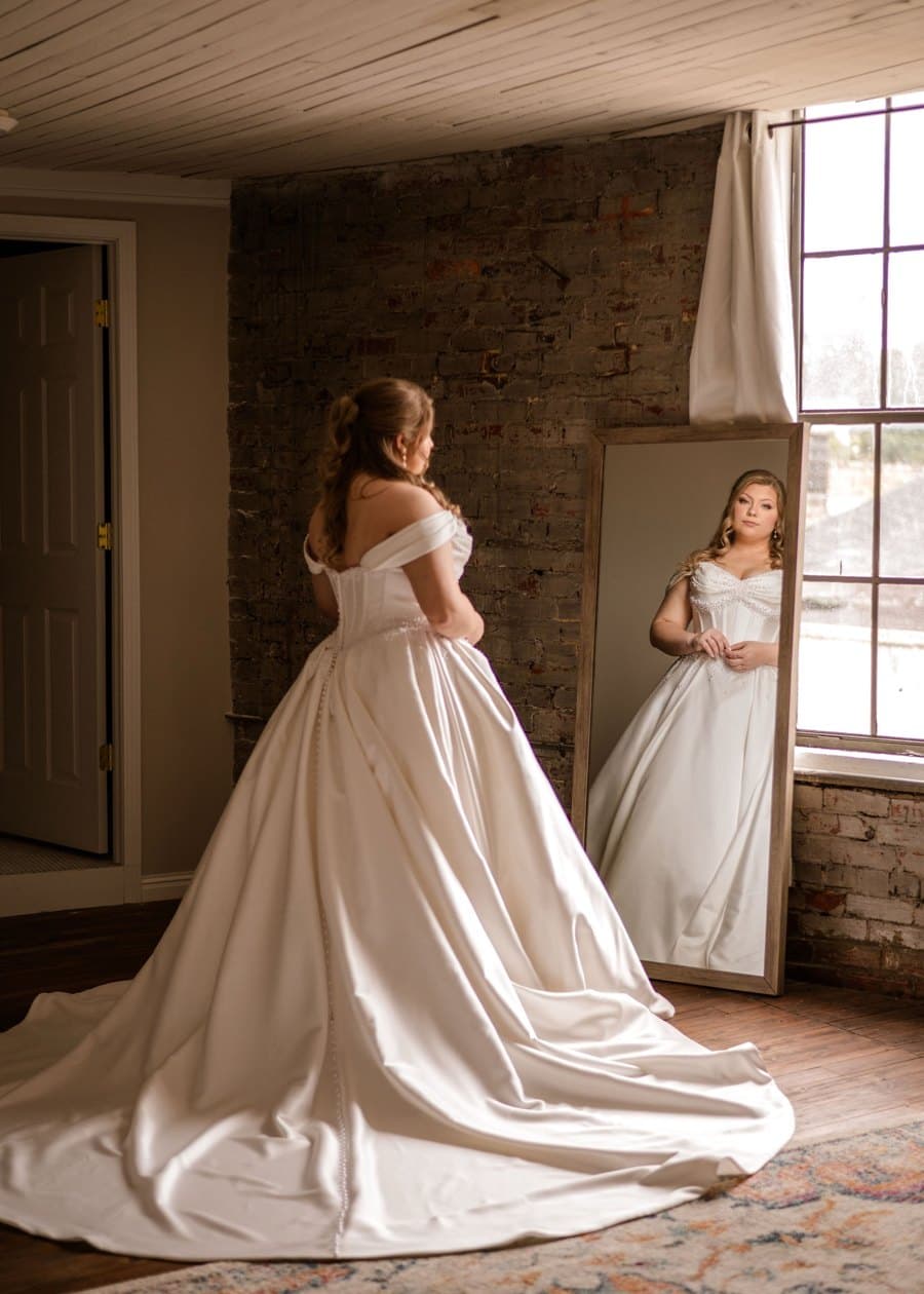 Bride in her gown looking into a tall mirror by the window