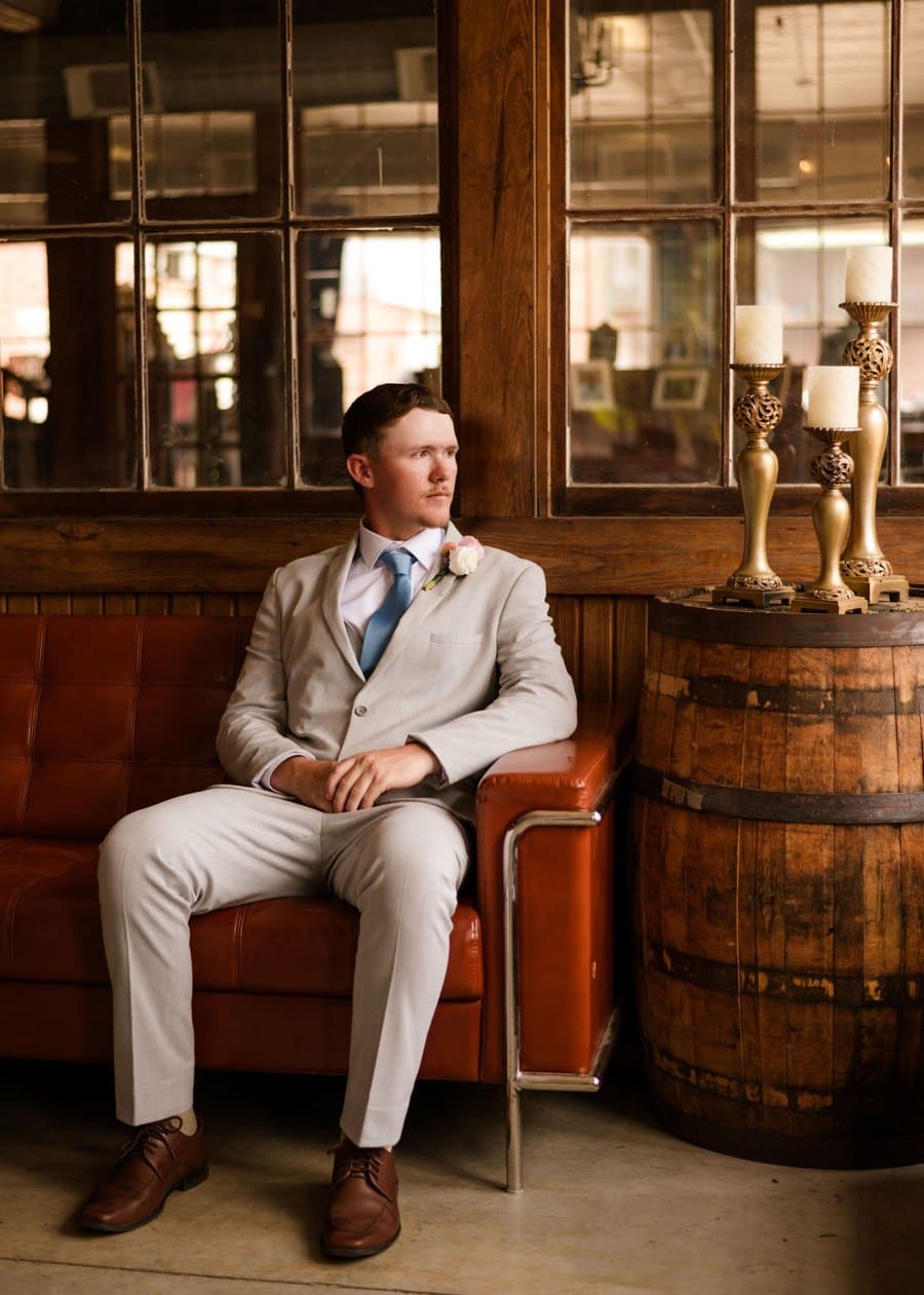 Groom in a light gray suit seated on a leather sofa by old wood-framed windows